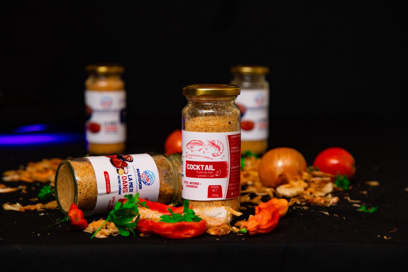 Pots d’assaisonnement MARIDOR Cocktail et La mer dans votre cuisine, entourés de piments, crevettes, tomates et oignon sur fond noir